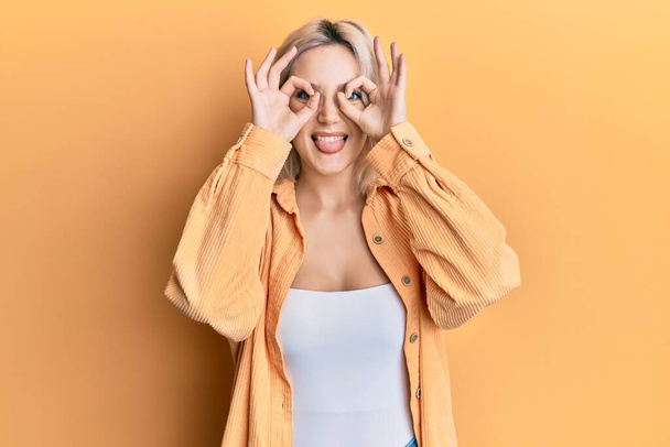 Young blonde girl wearing casual clothes doing ok gesture like binoculars sticking tongue out, eyes looking through fingers. crazy expression.  - Foto, Imagen