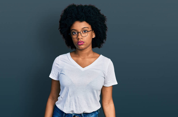 カジュアルな白いTシャツを着た若いアフリカ系アメリカ人女性が、真剣な表情でリラックスしています。カメラを見ているだけで.  - 写真・画像