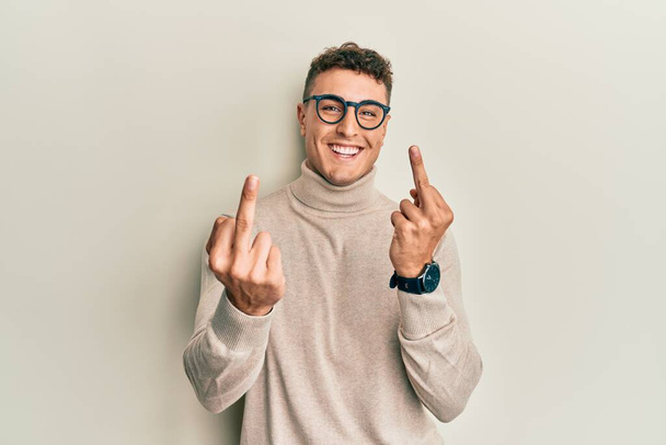 Hispanic young man wearing casual turtleneck sweater showing middle finger doing fuck you bad expression, provocation and rude attitude. screaming excited  - 写真・画像