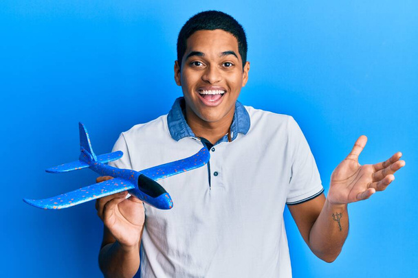 Young handsome hispanic man holding airplane toy celebrating achievement with happy smile and winner expression with raised hand  - 写真・画像