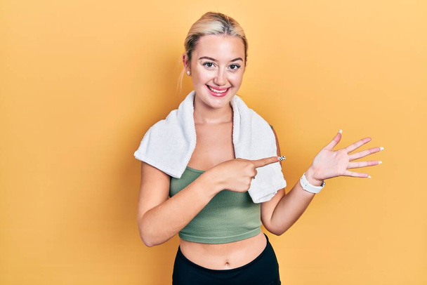 Молодая блондинка в спортивной одежде и полотенце удивлена и улыбается в камеру, представляя с рукой и указывая пальцем.  - Фото, изображение