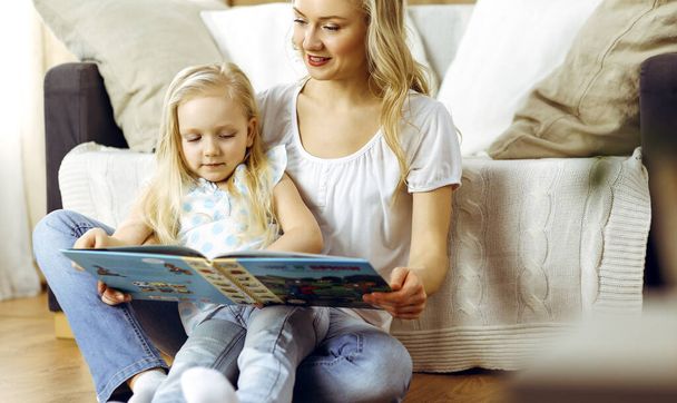 Happy family. Blonde young mother reading a book to her cute daughter while sitting at wooden floor. Motherhood concept - Photo, image