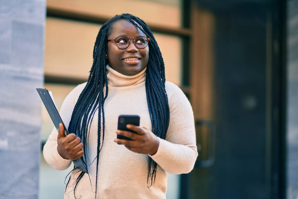 Nuori afrikkalainen amerikkalainen nainen, joka käyttää älypuhelinta ja pitää leikepöytää kaupungissa. - Valokuva, kuva