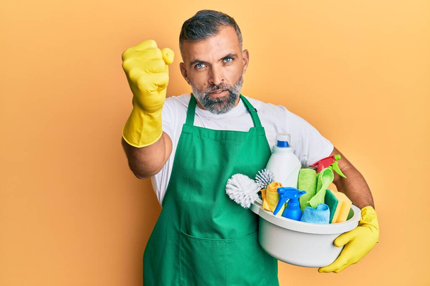 Homem bonito meia idade segurando produtos de limpeza irritado e frustrado gritando com raiva, gritando louco com raiva e mão levantada  - Foto, Imagem