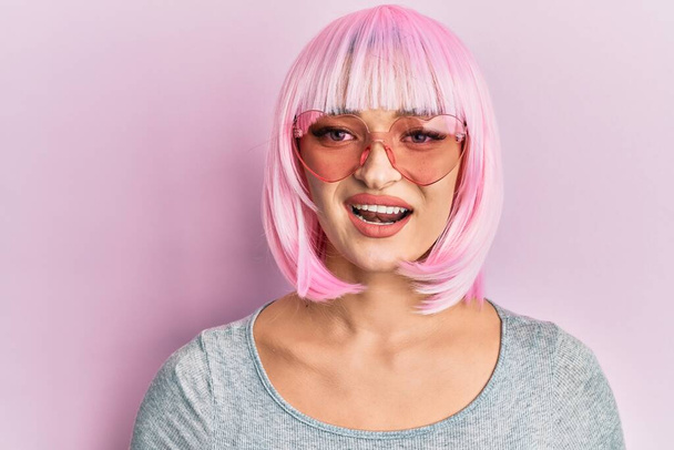 Młoda kaukaska kobieta ubrana w różową perukę i okulary przeciwsłoneczne serca wyglądająca pozytywnie i szczęśliwie stojąco i uśmiechnięta z pewnym siebie uśmiechem pokazującym zęby  - Zdjęcie, obraz