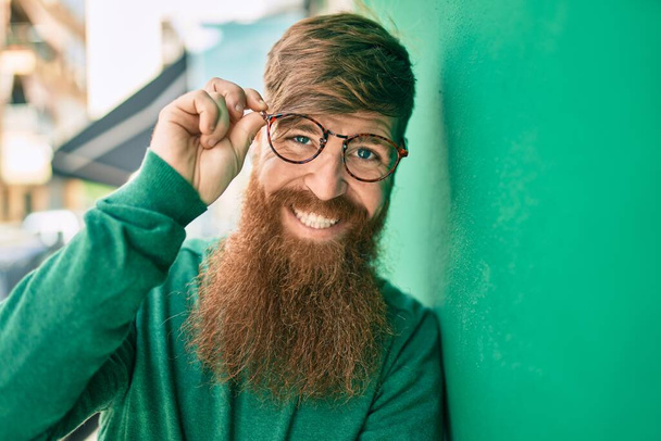 Fiatal ír férfi vörös szakállal mosolyogva boldogan hajol a falra a városban.. - Fotó, kép
