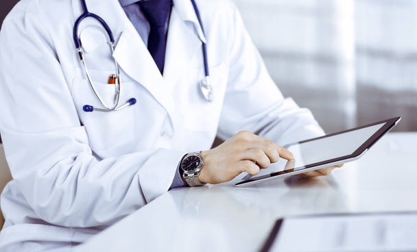 Unknown male doctor sitting and working with tablet computer in clinic at his working place, close-up. Young physician at work. Perfect medical service, medicine concept - Photo, Image