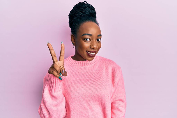 Young african american woman wearing casual winter sweater smiling looking to the camera showing fingers doing victory sign. number two.  - Photo, Image