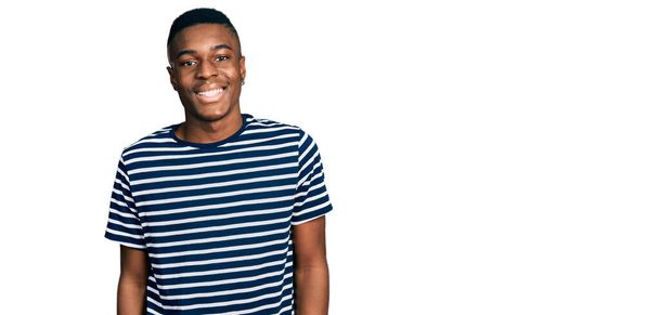 Молодой африканский американец в обычной полосатой футболке со счастливой и прохладной улыбкой на лице. счастливчик.  - Фото, изображение