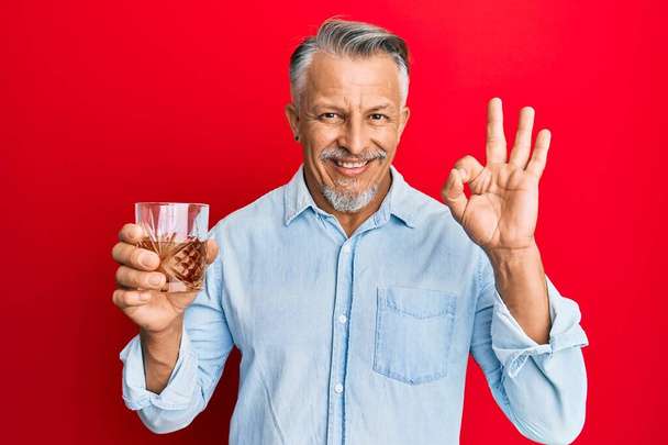 Middelbare leeftijd grijsharige man drinken glas whisky doet ok teken met de vingers, glimlachend vriendelijk gebaar uitstekend symbool  - Foto, afbeelding