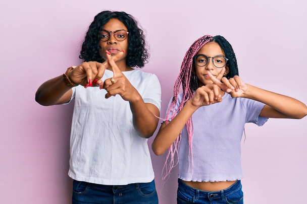 Hermosa madre afroamericana y su hija con ropa casual y gafas expresión de rechazo cruzando dedos haciendo signo negativo  - Foto, imagen