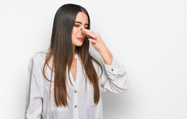 Young beautiful woman wearing casual white shirt smelling something stinky and disgusting, intolerable smell, holding breath with fingers on nose. bad smell  - Photo, Image