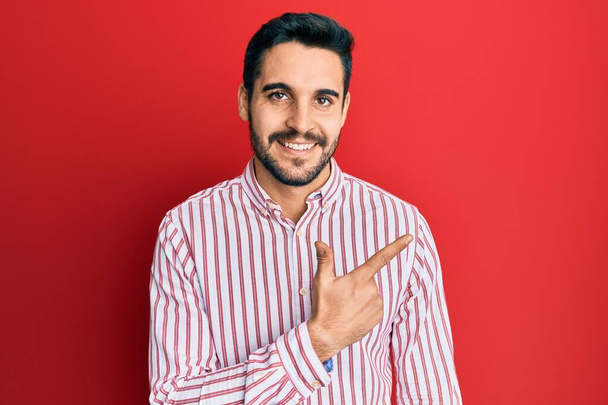 Νεαρός Ισπανός που φοράει επαγγελματική μπλούζα χαμογελώντας χαρούμενος δείχνοντας με το χέρι και το δάχτυλο στο πλάι  - Φωτογραφία, εικόνα