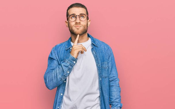 Junger kaukasischer Mann in lässiger Kleidung, der konzentriert mit dem Finger am Kinn über Zweifel nachdenkt und fragend nach oben schaut  - Foto, Bild