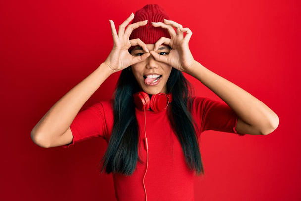 Giovane donna cinese che ascolta musica con le cuffie facendo gesto ok come binocolo sporgere la lingua, occhi che guardano attraverso le dita. espressione folle.  - Foto, immagini