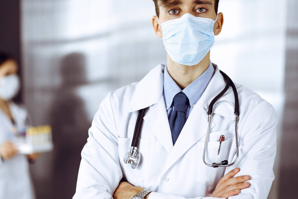 Young male doctor standing with arms crossed in clinic. Smart physician wearing medical protective mask in modern hospital. Medicine and Corona virus concept - Foto, Bild