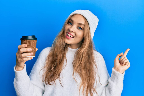 Schöne blonde kaukasische Frau trinkt eine Tasse Kaffee zum Mitnehmen und lächelt glücklich und zeigt mit Hand und Finger zur Seite  - Foto, Bild