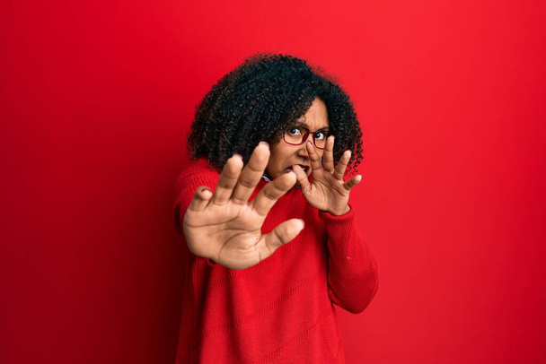 Mooie Afro-Amerikaanse vrouw met afro haar draagt trui en bril bang en doodsbang van angst uitdrukking stop gebaar met de handen, schreeuwen in shock. paniekconcept.  - Foto, afbeelding