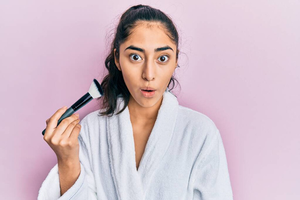 Hispanic teenager girl with dental braces wearing robe holding makeup brush scared and amazed with open mouth for surprise, disbelief face  - Foto, Imagen