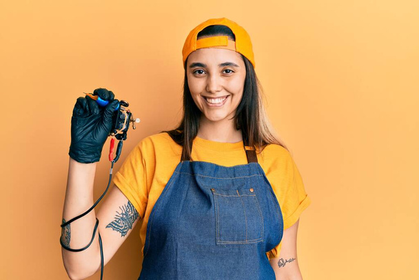 Junge hispanische Tätowiererin trägt professionelle Uniform und Handschuhe, die Tätowierer Maschine sieht positiv und glücklich stehend und lächelnd mit einem selbstbewussten Lächeln Zähne zeigen  - Foto, Bild