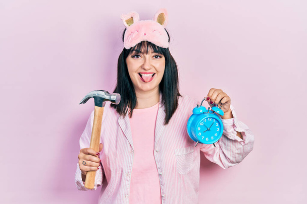 Young hispanic woman wearing sleep mask and robe holding alarm clock and hammer sticking tongue out happy with funny expression.  - Photo, Image