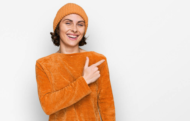 Mujer hispana joven vistiendo ropa casual y gorra de lana alegre con una sonrisa de la cara señalando con la mano y el dedo hacia el lado con expresión feliz y natural en la cara  - Foto, imagen