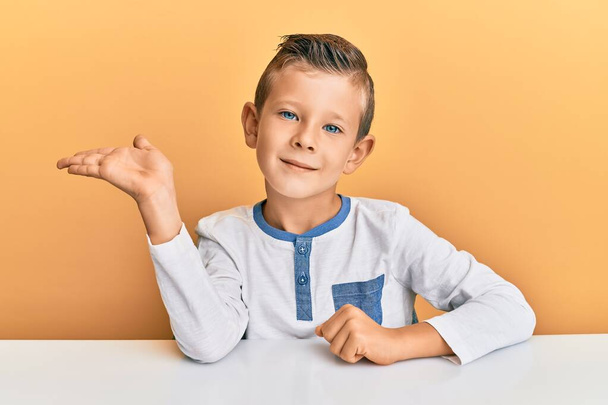 Adorable caucasian kid wearing casual clothes sitting on the table smiling cheerful presenting and pointing with palm of hand looking at the camera.  - Foto, immagini
