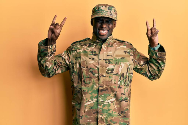 Young african american man wearing army uniform shouting with crazy expression doing rock symbol with hands up. music star. heavy music concept.  - Фото, изображение