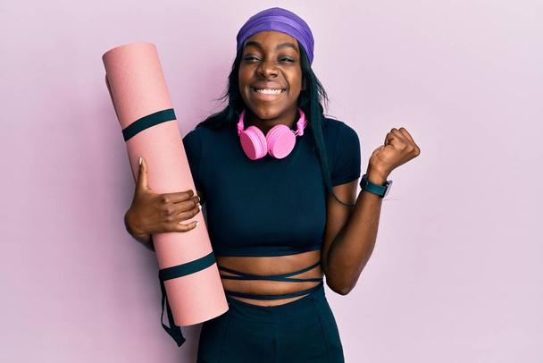 Junge Afroamerikanerin in Turnbekleidung hält Yogamatte schreiend stolz in der Hand und feiert Sieg und Erfolg sehr aufgeregt mit erhobenem Arm  - Foto, Bild