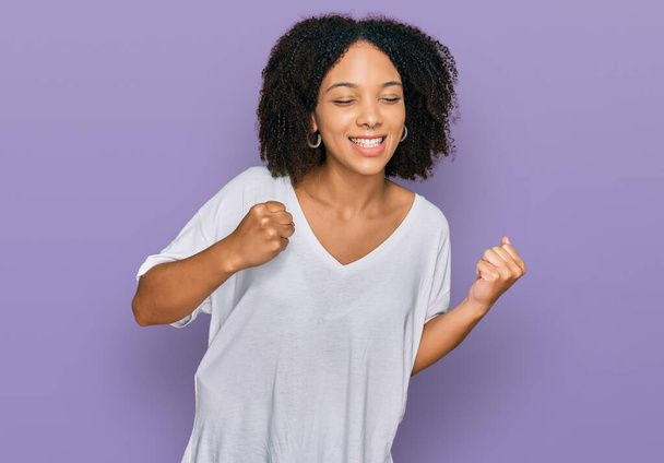 Nuori afrikkalainen amerikkalainen tyttö yllään rento vaatteita innoissaan menestykseen kädet ylhäällä ja silmät kiinni juhlii voittoa hymyillen. voittajan käsite.  - Valokuva, kuva