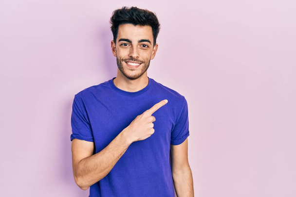 Joven hombre hispano vistiendo camiseta casual alegre con una sonrisa en la cara apuntando con la mano y el dedo hacia un lado con expresión feliz y natural  - Foto, Imagen