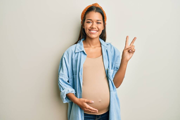 Mooie Latijns-Amerikaanse vrouw die een baby verwacht, die een zwangere buik aanraakt en glimlacht terwijl ze naar de camera kijkt en vingers laat zien die een overwinningsteken doen. nummer twee.  - Foto, afbeelding
