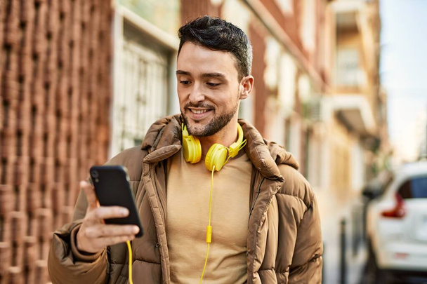 Молодий іспаномовний чоловік посміхається щасливим, використовуючи смартфон та навушники у місті
. - Фото, зображення