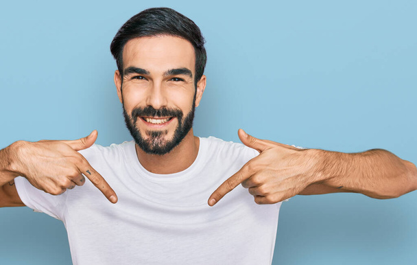 Fiatal spanyol férfi alkalmi fehér pólót visel, magabiztos mosollyal az arcán, ujjaival büszkén és boldogan mutogat magára..  - Fotó, kép