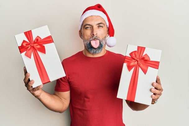 Middle age handsome man wearing christmas hat and holding a gifts sticking tongue out happy with funny expression.  - Foto, Bild