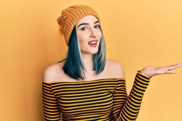 jong modern meisje dragen wol hoed lachende vrolijke presenteren en wijzen met palm van de hand kijken naar de camera.  - Foto, afbeelding