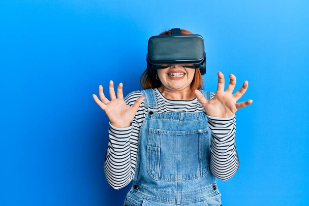 Fiatal vörös hajú nő megijedt segítségével 3D virtuális szemüveg áll elszigetelt kék háttér. - Fotó, kép