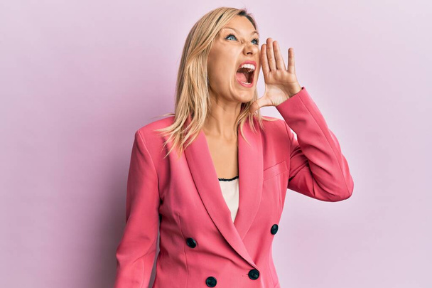 Mujer caucásica de mediana edad con chaqueta de negocios gritando y gritando fuerte a un lado con la mano en la boca. concepto de comunicación.  - Foto, Imagen