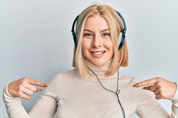 Молода блондинка слухає музику, використовуючи навушники, які виглядають впевнено з посмішкою на обличчі, вказуючи на себе з пальцями гордими і щасливими
.  - Фото, зображення