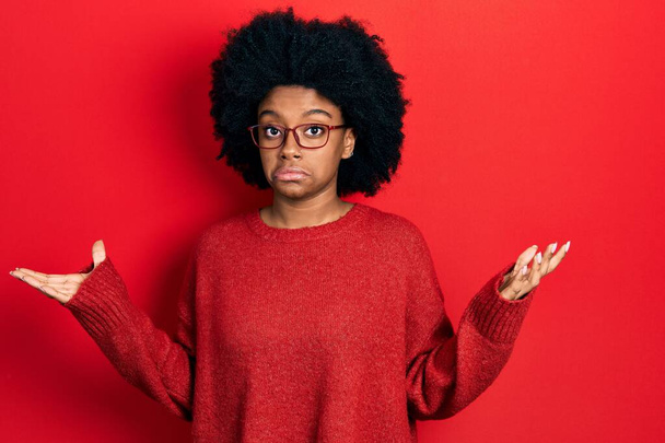 Jeune femme afro-américaine portant des vêtements et des lunettes décontractés sans savoir et confus avec les bras ouverts, aucune idée et le visage douteux.  - Photo, image