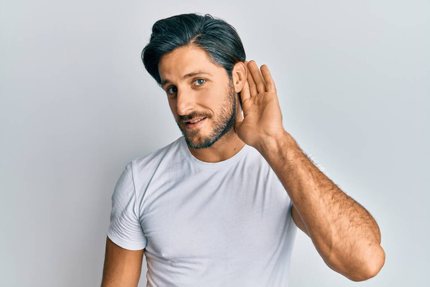 Young hispanic man wearing casual white t shirt smiling with hand over ear listening and hearing to rumor or gossip. deafness concept.  - Photo, Image