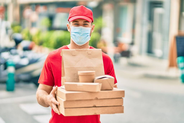 Joven repartidor caucásico con máscara médica sosteniendo comida a domicilio en la ciudad. - Foto, imagen