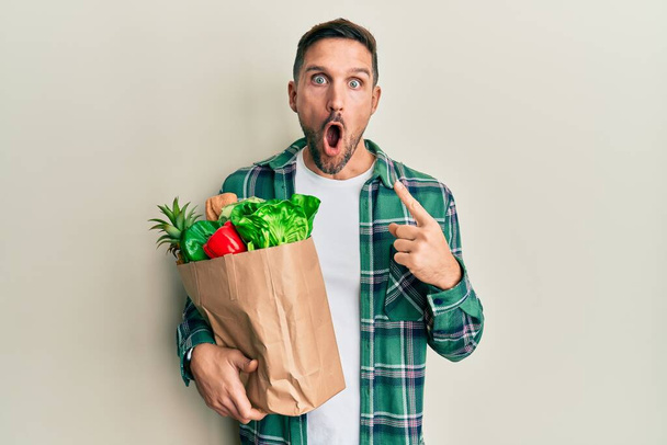 Pohledný muž s plnovousem, držící papírový sáček s potravinami, překvapený ukazováčkem na stranu, s otevřenými ústy užaslý výraz.  - Fotografie, Obrázek
