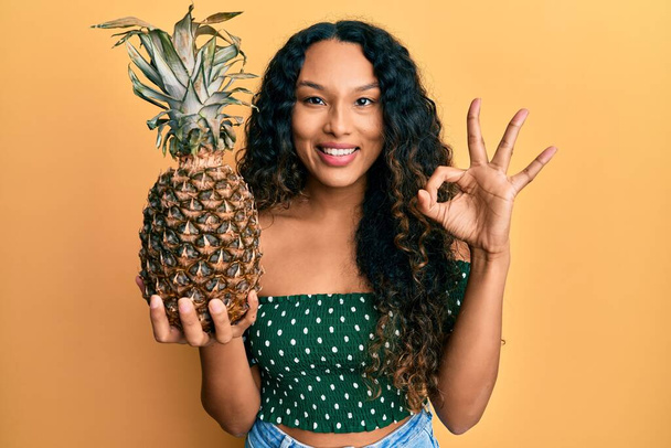Fiatal latin nő kezében ananász csinál rendben jel ujjaival, mosolygós barátságos gesztus kiváló szimbólum  - Fotó, kép