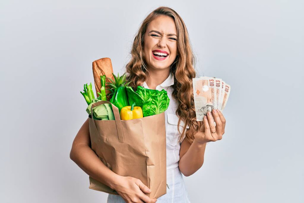 Young blonde girl holding paper bag with groceries and pounds smiling and laughing hard out loud because funny crazy joke.  - 写真・画像