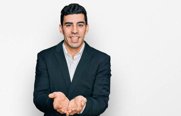 Hombre hispano guapo vistiendo ropa de negocios sonriendo con las palmas de las manos juntas recibiendo o dando gesto. retención y protección  - Foto, Imagen