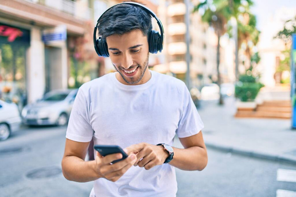 Νεαρός Λατίνος άνθρωπος χαμογελώντας χαρούμενος χρησιμοποιώντας smartphone και ακουστικά στην πόλη. - Φωτογραφία, εικόνα
