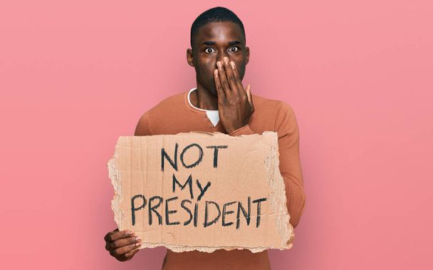 Junger afrikanisch-amerikanischer Mann, der nicht das Protestbanner meines Präsidenten in der Hand hält, das den Mund mit der Hand bedeckt, schockiert und Angst vor Fehlern hat. Überraschte Miene  - Foto, Bild