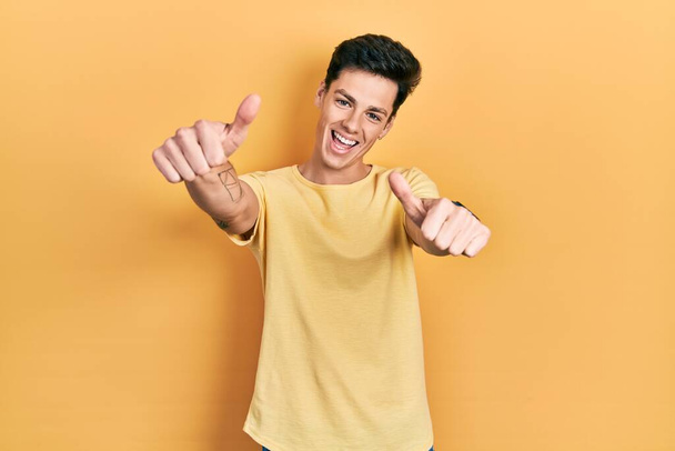 Junger hispanischer Mann in lässigem, gelbem T-Shirt, der zustimmend positive Gesten mit der Hand macht, Daumen hoch lächelnd und glücklich über den Erfolg. Siegergeste.  - Foto, Bild