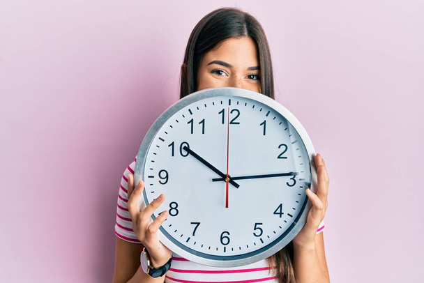 Young brunette woman holding big clock covering face smiling with a happy and cool smile on face. showing teeth.  - Zdjęcie, obraz